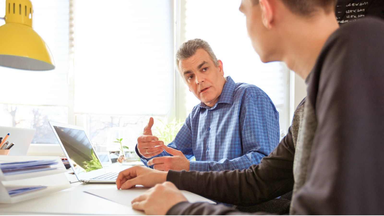 A man in his 60s, wearing a blue shirt, talks to a teenage boy wearing dark clothes. A laptop and desk is visible.
