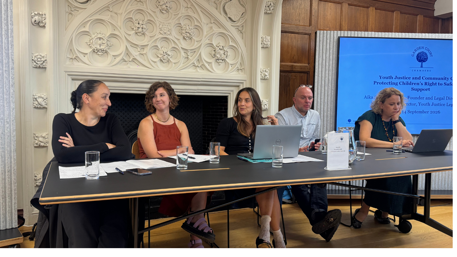 All the panel members sitting behind a desk with a PowerPoint presentation in the background